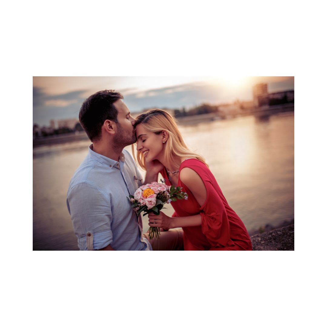 A couple embracing each other by a body of water with a sunset in the background, the woman holding a bouquet of flowers.