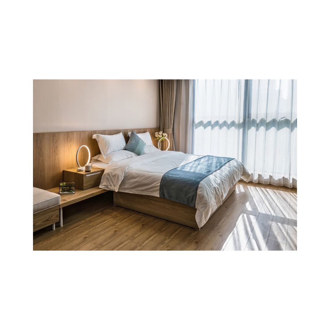 A well-lit, tidy bedroom with a large bed, wooden headboard, and a nightstand with a lamp and a book on it. The room has a modern aesthetic with a bright window and a balcony.