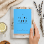 Book titled 'The Clear Path' on a white surface with a hand holding it, surrounded by a cup of tea, lemon, and lavender.