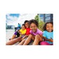 A group of four smiling children sitting close by a body of water, with a fifth child standing behind them. They are wearing colorful clothing.